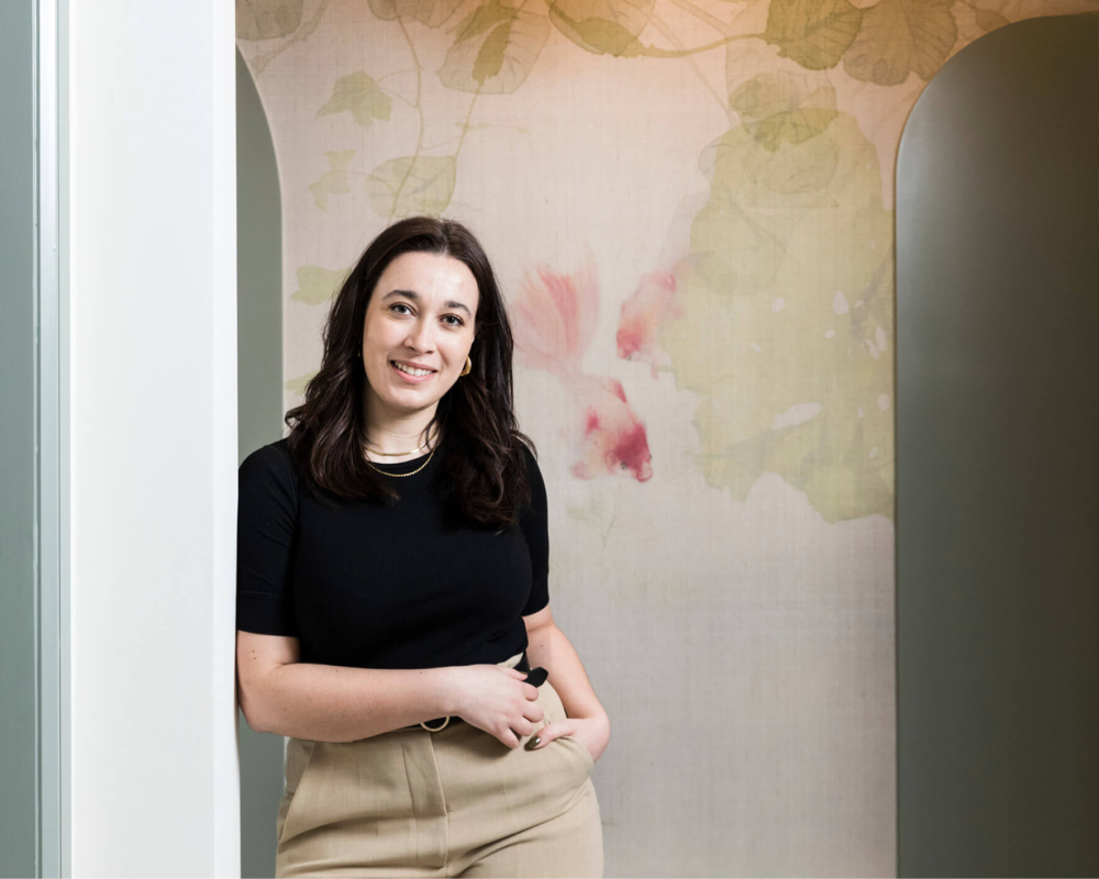 Becky from the patient services team is standing by a wall smiling to the camera