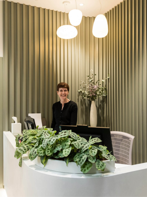 Image of the reception desk covered in computers and plants with the receptionist standing up at the Evewell fertility clinic