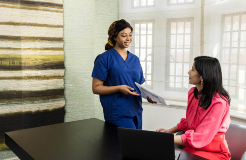 Nurse standing up handing patient fertility files to a consultant who is sitting on a desk with her laptop