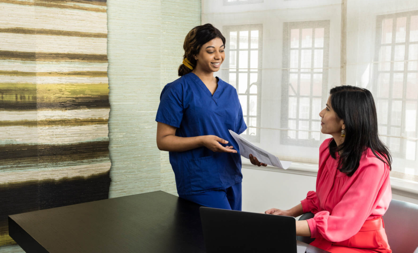 Nurse standing up handing patient fertility files to a consultant who is sitting on a desk with her laptop